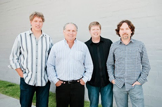 Four men of varying ages standing side-by-side in casual button-down shirts against a gray wall.