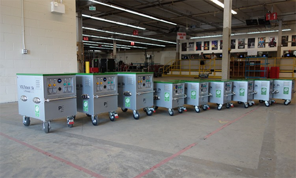 A line of silver metal mobile power units with green accents on wheels in a large warehouse.
