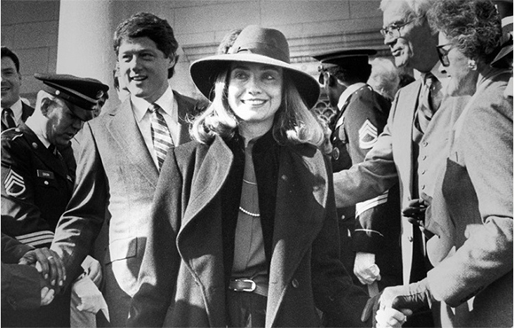 A smiling woman in a dark coat and wide-brimmed hat walks through a crowd of people.