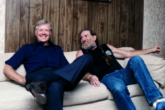 Jimmy Carter and a man with long hair and a beard sitting on a beige couch against wood paneling.
