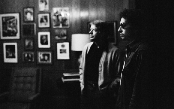 Black and white profile shot of two men standing in a room filled with framed photographs.