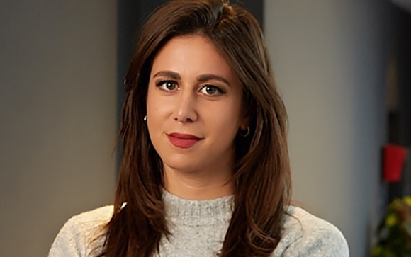 A woman with long brown hair and dark eyes wearing a grey sweater looks toward the camera.