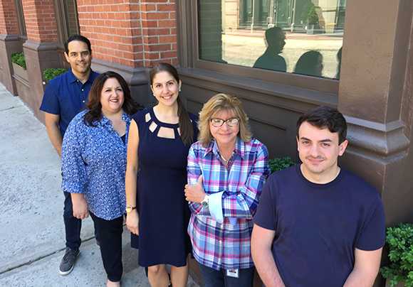 Five people standing in a row on a sidewalk in front of a brick building and large window.