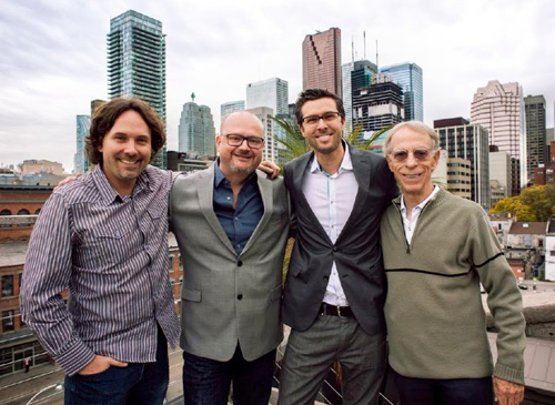 Four men smiling and posing together on a rooftop with a city skyline in the background.