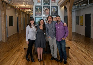 Four people stand together in a hallway with wooden floors and movie posters on the wall.
