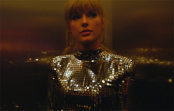 A woman with blonde hair wearing a silver sequined high-neck top against a dark, blurred background.