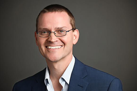 A smiling man with short brown hair and glasses wearing a blue suit jacket and light blue shirt.