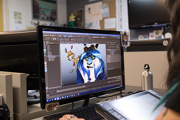 A person uses a digital tablet to edit a blue animated character on a computer screen.