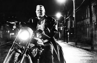 A man in a leather jacket and white shirt rides a motorcycle down a street at night under streetlights.