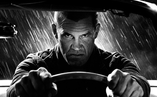 Black and white close-up of a man with a serious expression gripping a steering wheel in heavy rain.