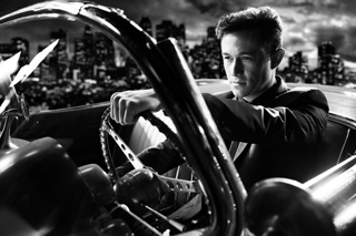 Black and white shot of a man in a suit driving a classic convertible through a city skyline at night.