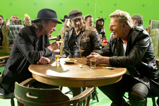 Several men sit around a wooden table with drinks in a room featuring a bright green screen background.