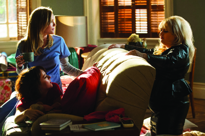 A woman in blue scrubs holds a mug while another woman in black leans over a sofa near a woman lying down.