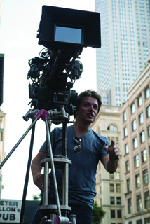 A man gestures while standing beside a large professional film camera mounted on a tripod in a city.