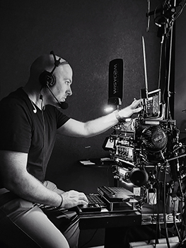 A man wearing a headset operates a complex array of audio and video equipment in a dark studio.
