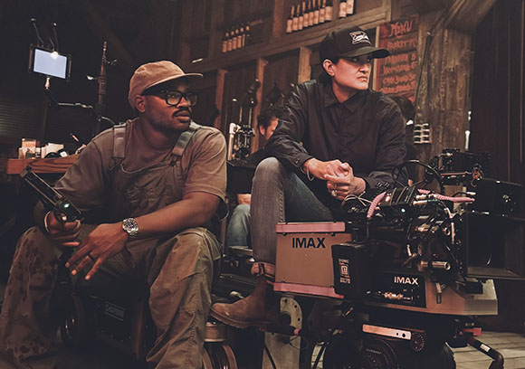 Two crew members sit behind a large IMAX camera in a dimly lit, rustic indoor set.
