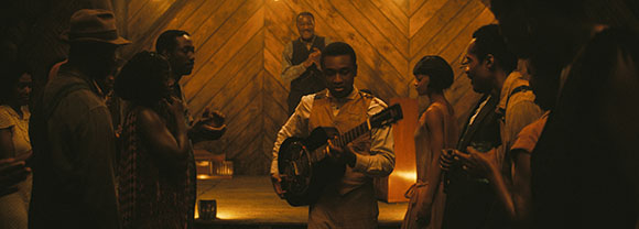 A man plays an acoustic guitar in a warm, dimly lit room surrounded by a group of people.