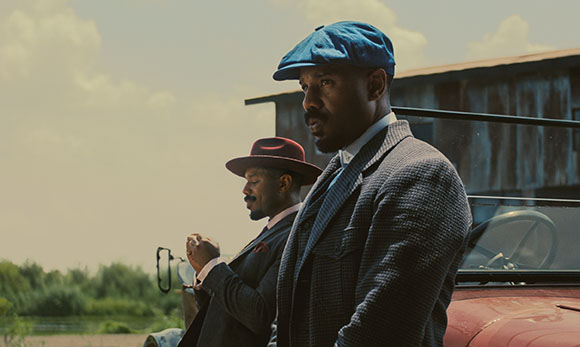 Two men in suits and hats stand outdoors near a vintage vehicle under a cloudy sky.