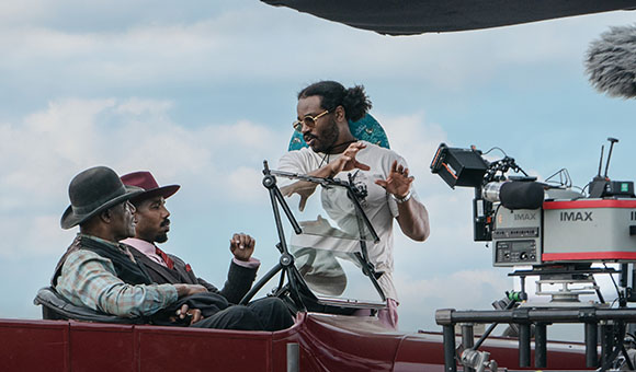 A director in sunglasses gestures toward two seated actors on a film set under a cloudy sky.
