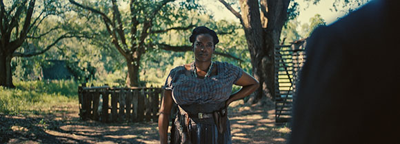 A woman in period clothing stands with hands on hips in a sunlit, wooded clearing.