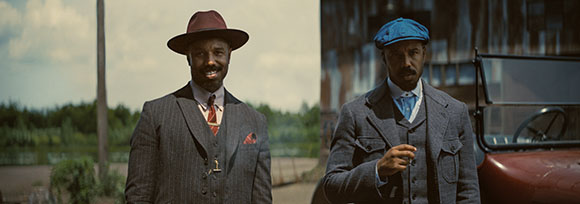 Two men wearing vintage suits and hats standing outdoors near a car.