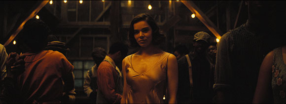 A woman in a gold sleeveless dress looks toward the camera in a crowded, dimly lit room with warm string lights.