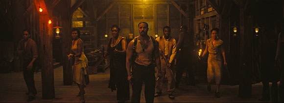 A group of people stand in a dimly lit, rustic wooden interior with warm orange lighting.
