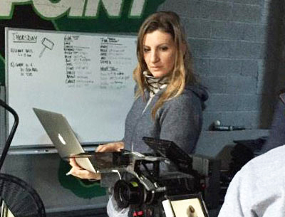 A woman in a blue hoodie looks at an open laptop in a workspace with a whiteboard behind her.