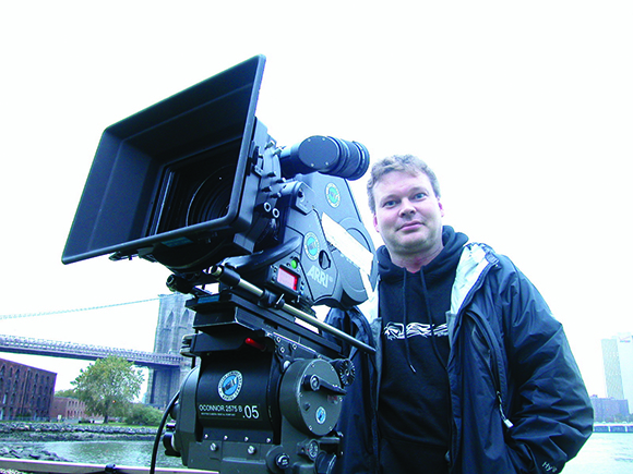 A man in a dark jacket stands beside a large professional film camera on a tripod.