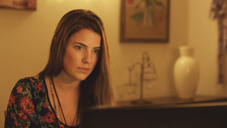 A young woman with long brown hair looks intently at a computer screen in a dimly lit room.