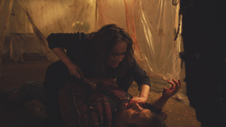 A woman in dark clothing leans over a man lying on the ground in a dark, cluttered room.