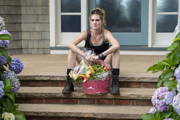 A woman in black clothing sits on stone steps behind a large pink basket filled with vegetables.