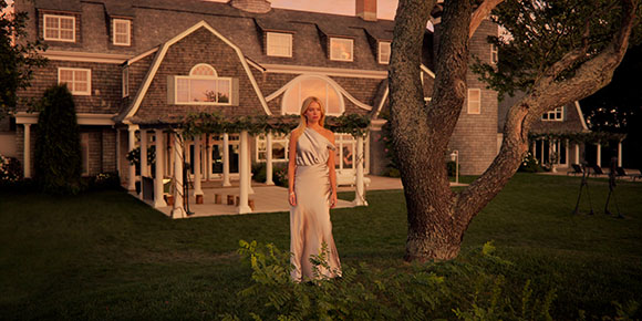 A woman in a long, one-shoulder light gown stands on a lawn before a large, lit house at twilight.