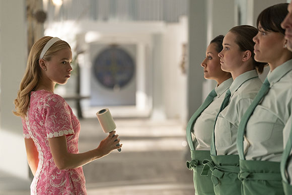 A blonde woman in a pink patterned dress holds a hair roller while facing four women in light green uniforms.