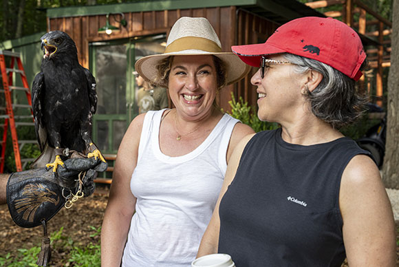 A woman in a white tank top smiles next to a person holding a large dark bird of prey.