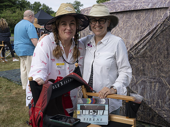 Two women in sun hats smile behind a film clapperboard labeled 'SIRENS' on an outdoor set.