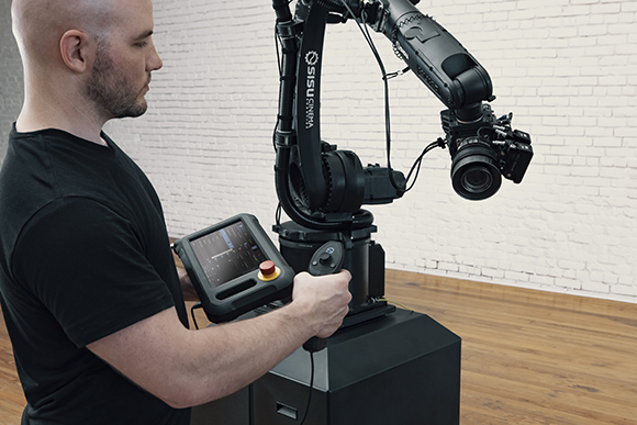 A man in a black shirt uses a handheld control panel to operate a large black robotic arm holding a camera.