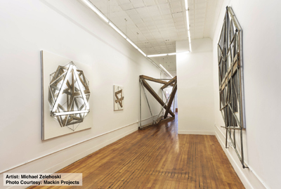 A long white gallery with wooden floors displaying various geometric metal and wood sculptures on the walls.