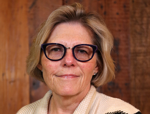 A woman with blonde hair and dark-rimmed glasses smiling slightly against a wooden background.