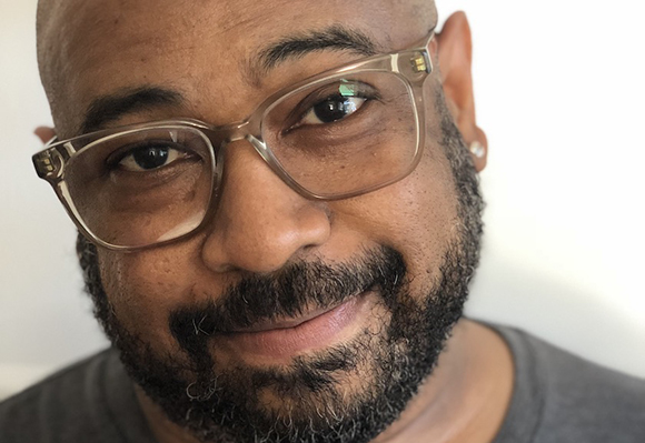 Close-up portrait of a man with glasses, a dark beard, and a slight smile against a white background.