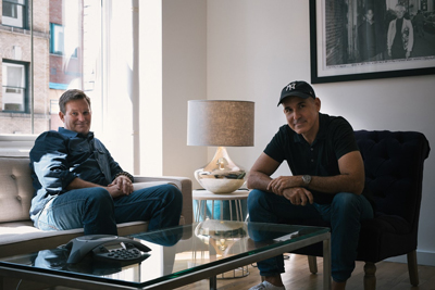Two men sit in a bright living room, one on a sofa and one in an armchair near a lamp and framed art.