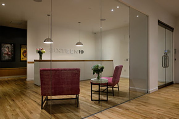 A reception desk with the text SIXTEEN19 on the wall, two pink chairs, and a small table behind a glass wall.