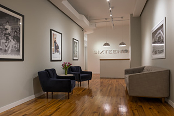 A lobby with hardwood floors, two blue velvet armchairs, a grey sofa, and framed art on the walls.