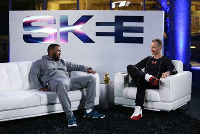 Two men sit on a white sofa and armchair facing each other in a studio with an SKEE logo background.