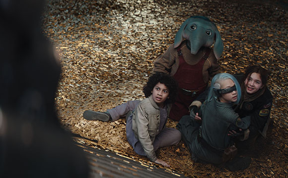 Four children sit on a floor covered in gold flakes, including one wearing an elephant mask.