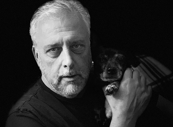 Black and white portrait of a man with gray hair and beard holding a small dog close to his face.
