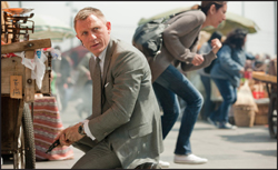 A man in a grey suit crouches low while a woman runs past him in a busy outdoor market.