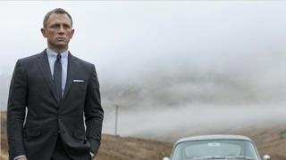 A man in a dark suit and tie stands in a misty, hilly landscape near a vintage car.