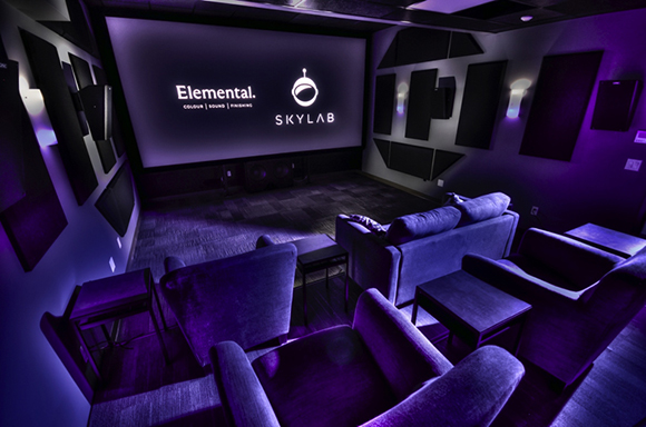 A dark movie screening room with purple ambient lighting, plush velvet sofas, and a large projection screen.