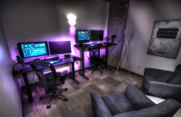 Two computer desks with monitors and ergonomic chairs next to two grey armchairs in a dimly lit room.